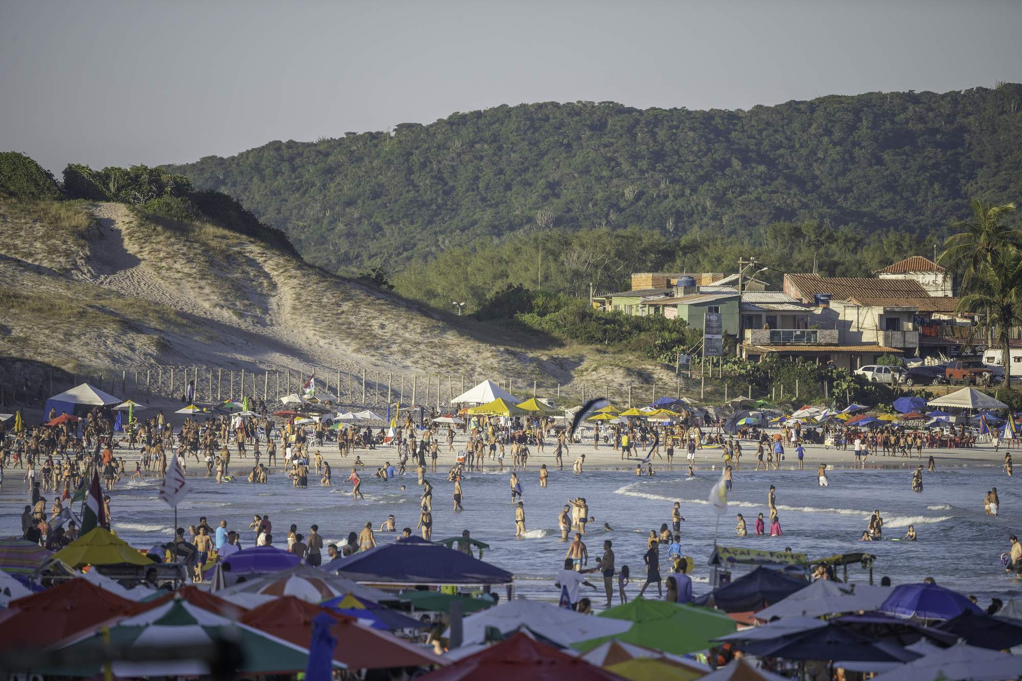 Praia do Forte lotada, em Cabo Frio, com guarda-sóis coloridos e dunas.