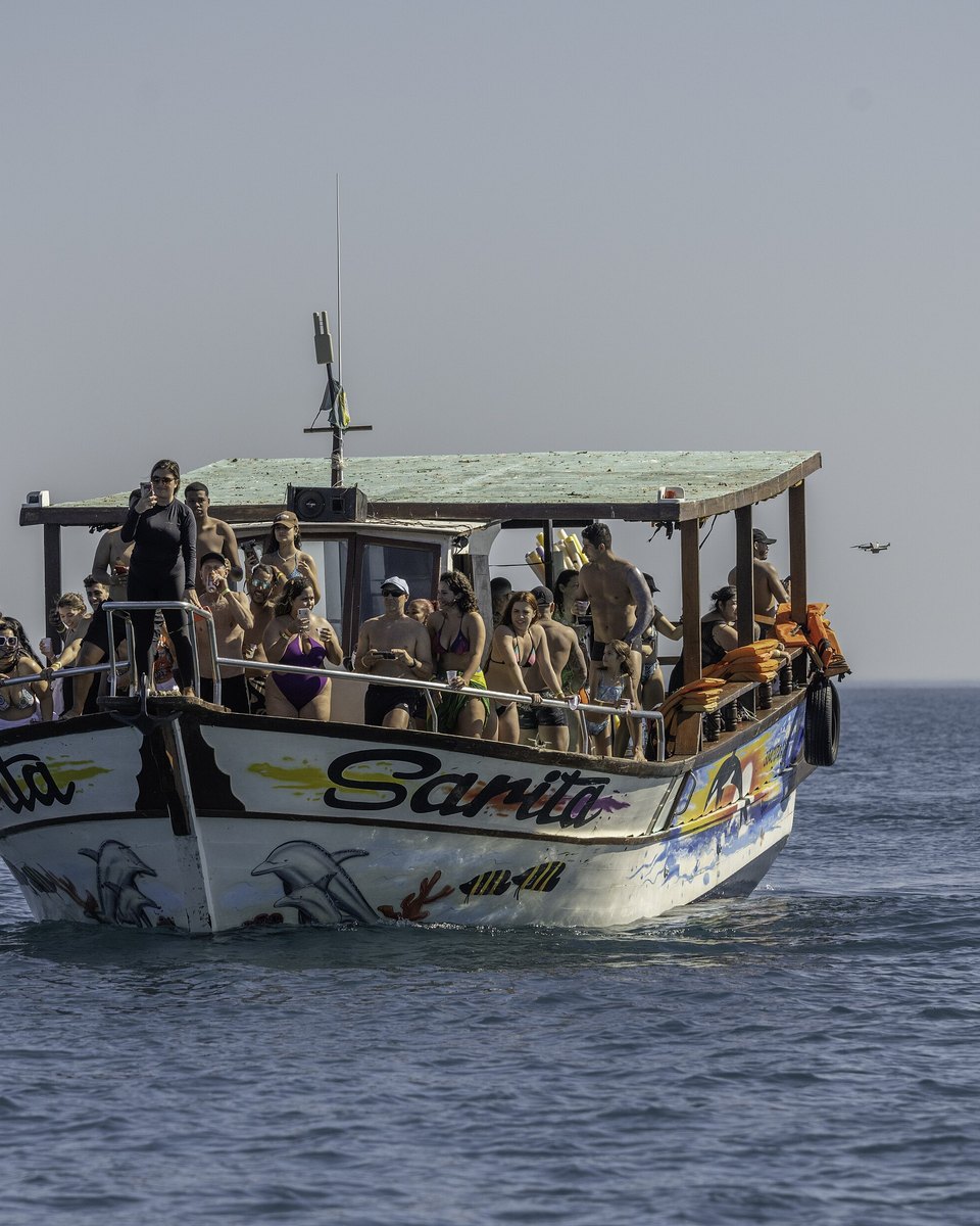 Barco de passeio de dois andares lotado de turistas de roupa de banho e colete salva-vidas laranja, navegando no mar calmo de Arraial do Cabo.
