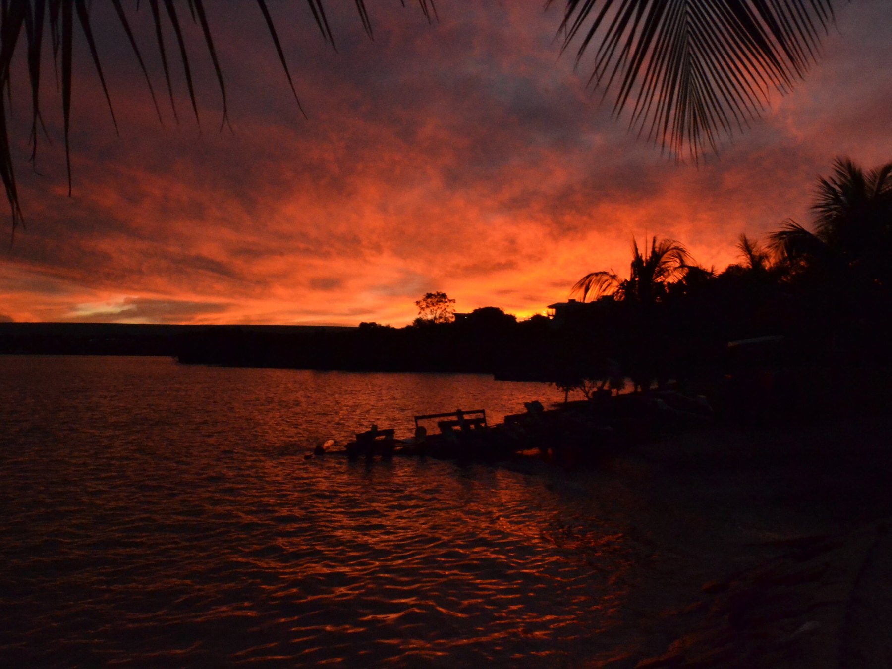 Pôr do sol no Lago Corumbá, com o céu vermelho e laranja refletido na água, folhas de coqueiro em primeiro plano e a silhueta da margem, de um píer e das árvores.