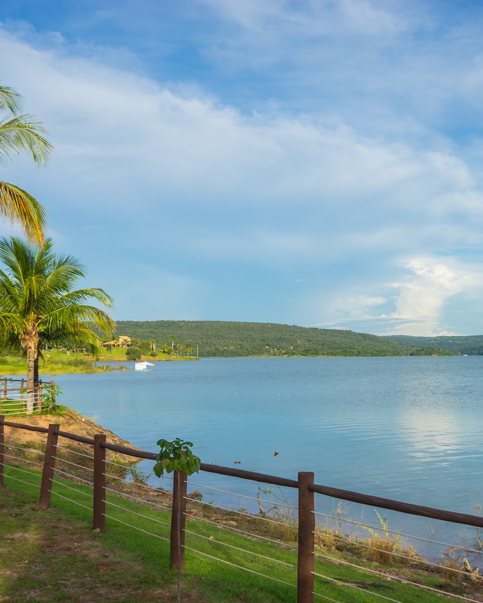 Margem gramada do Lago Corumbá, em Caldas Novas, com coqueiros, uma cerca de madeira na beira da água e o lago largo e azul se estendendo até os morros no horizonte, sob céu com nuvens.