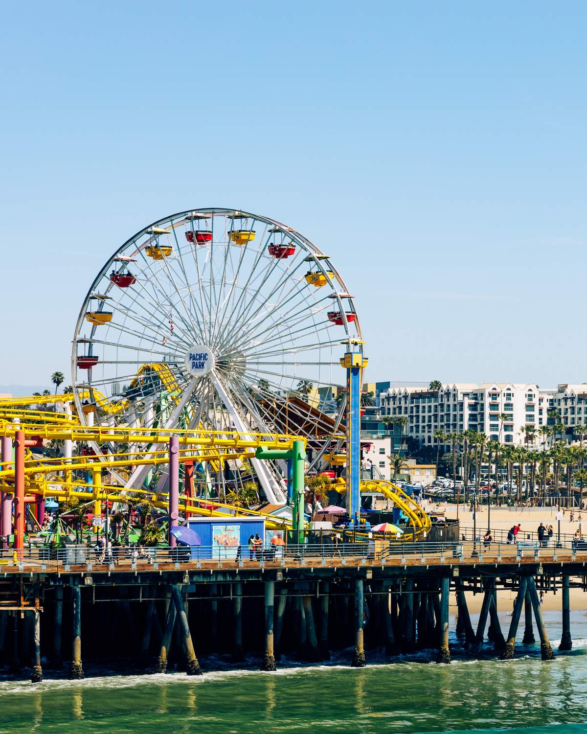 Roda-gigante e montanha-russa do parque do Santa Monica Pier sobre o mar, com a praia cheia de gente e os prédios de Santa Monica ao fundo.