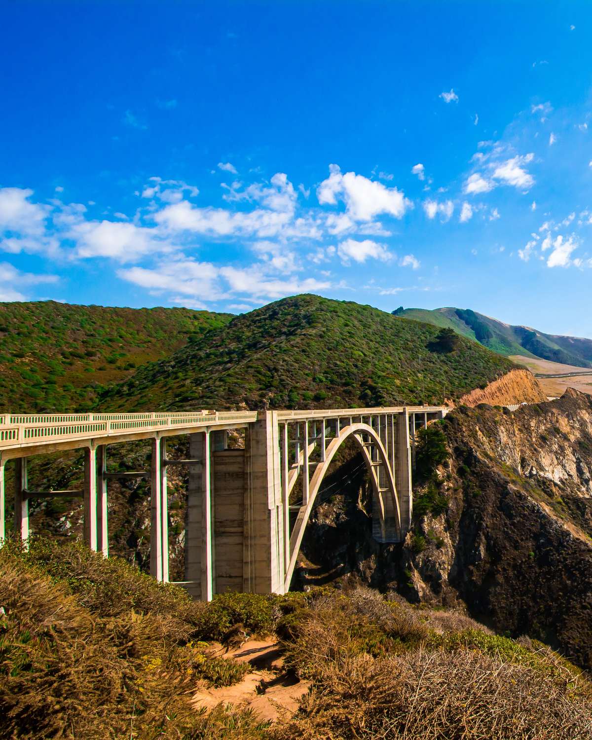 Ponte Bixby Creek, em arco de concreto, ligando dois penhascos verdes na Highway 1, com o Pacífico azul ao fundo.