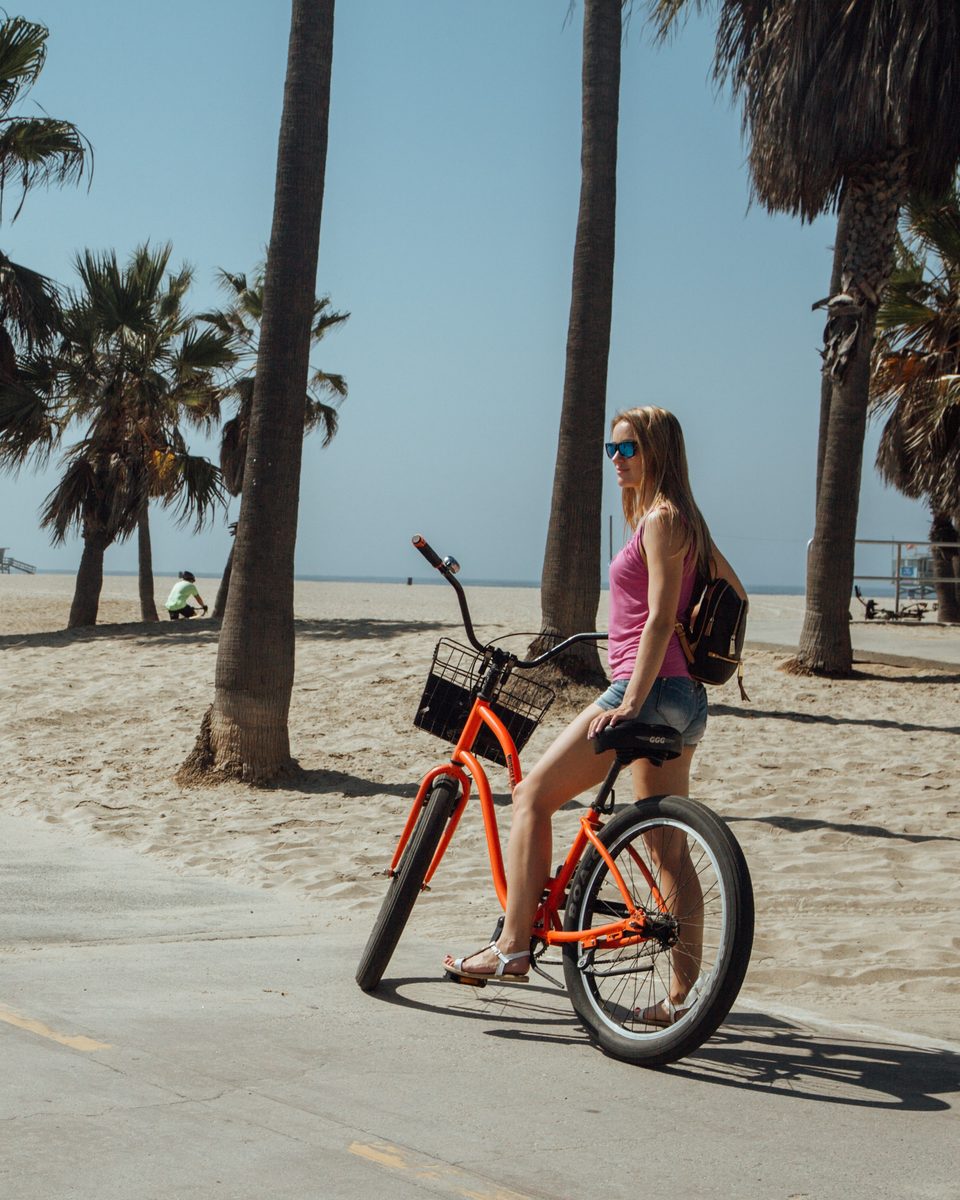 Mulher de bicicleta laranja parada na ciclovia à beira da areia em Venice Beach, entre palmeiras, num dia de sol.