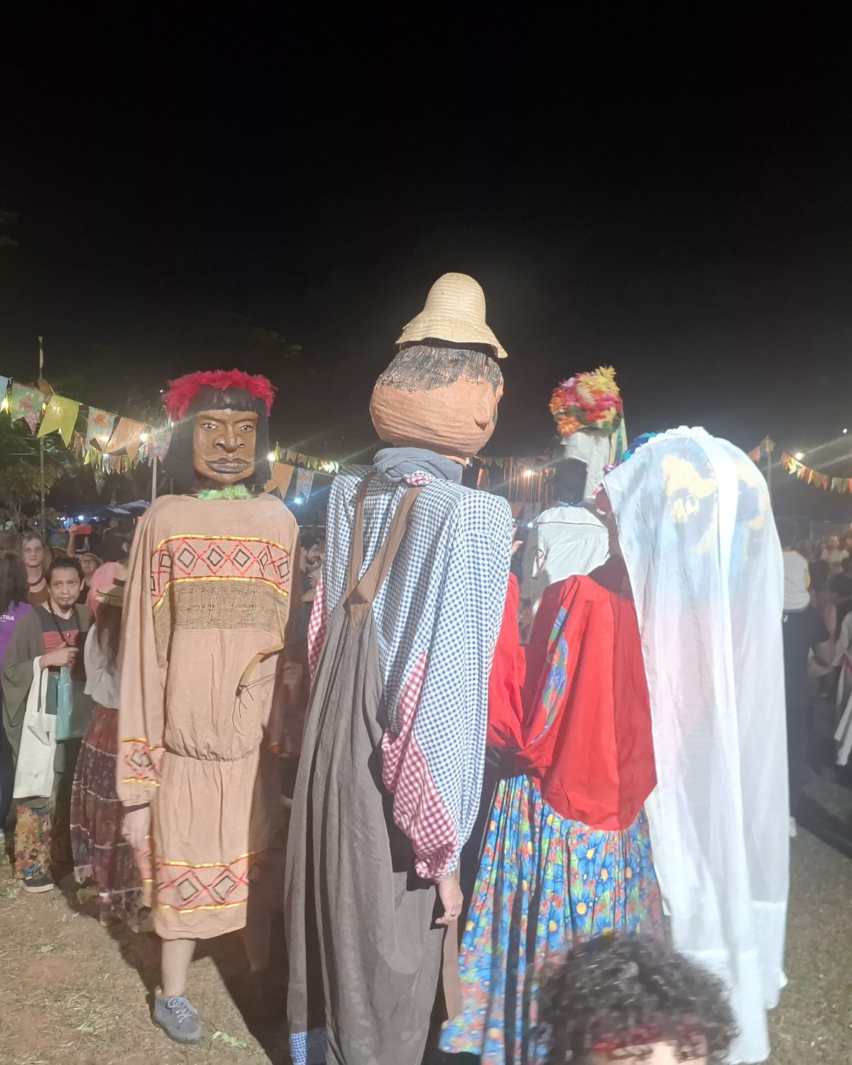 Bonecões gigantes no meio do público de uma festa junina à noite em Barão Geraldo, em Campinas, com bandeirinhas ao fundo.