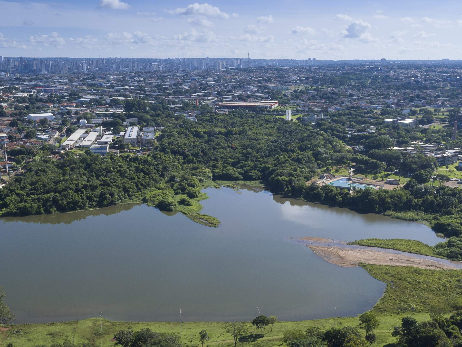 Vista aérea do Lago do Amor, em Campo Grande, com a água espelhada cercada de mata verde e os prédios da cidade no horizonte sob céu azul com nuvens