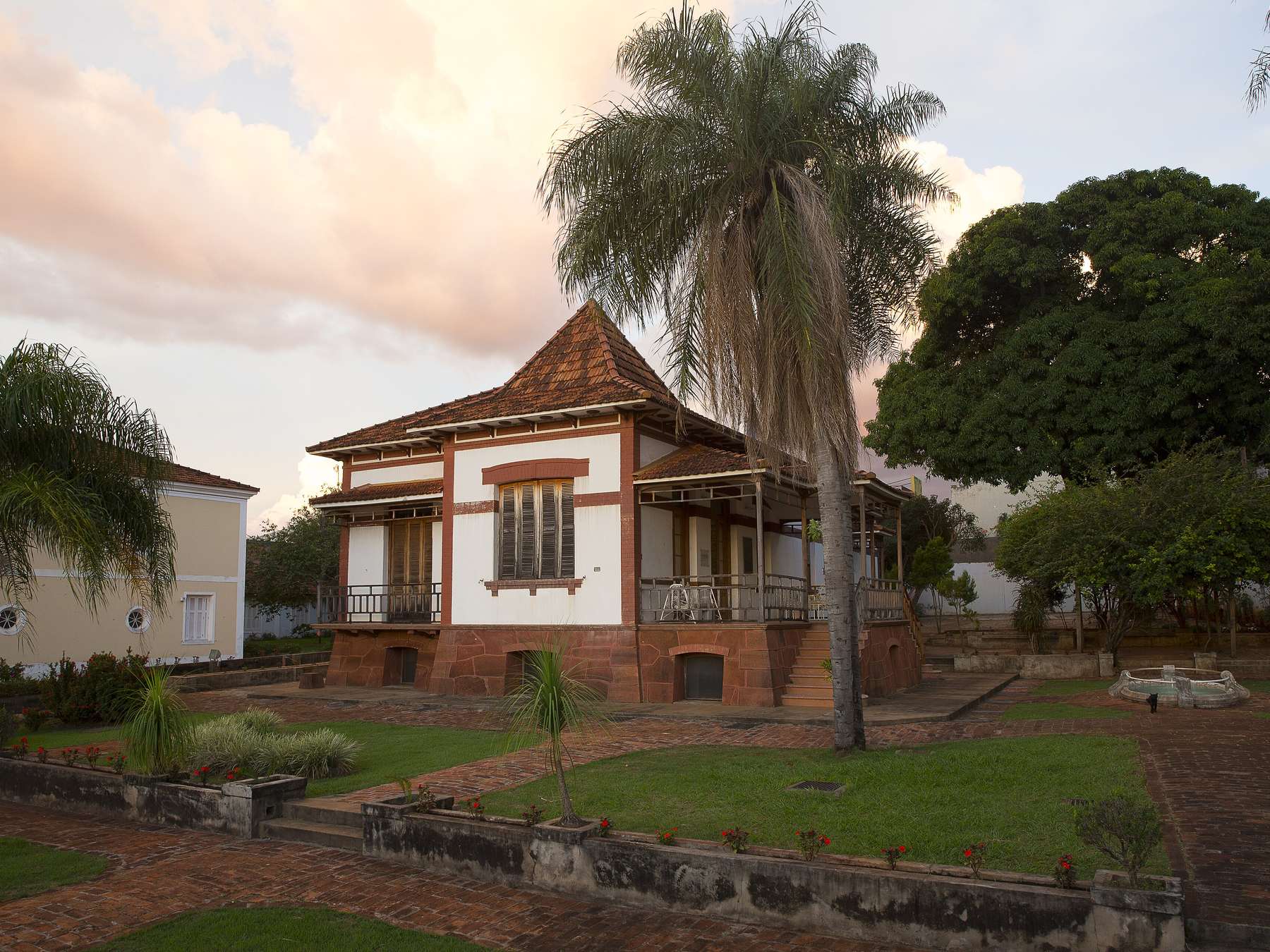 Casarão branco com detalhes em vermelho e varanda de ferro, cercado por palmeiras e jardim, iluminado pela luz rosada do fim de tarde em Campo Grande