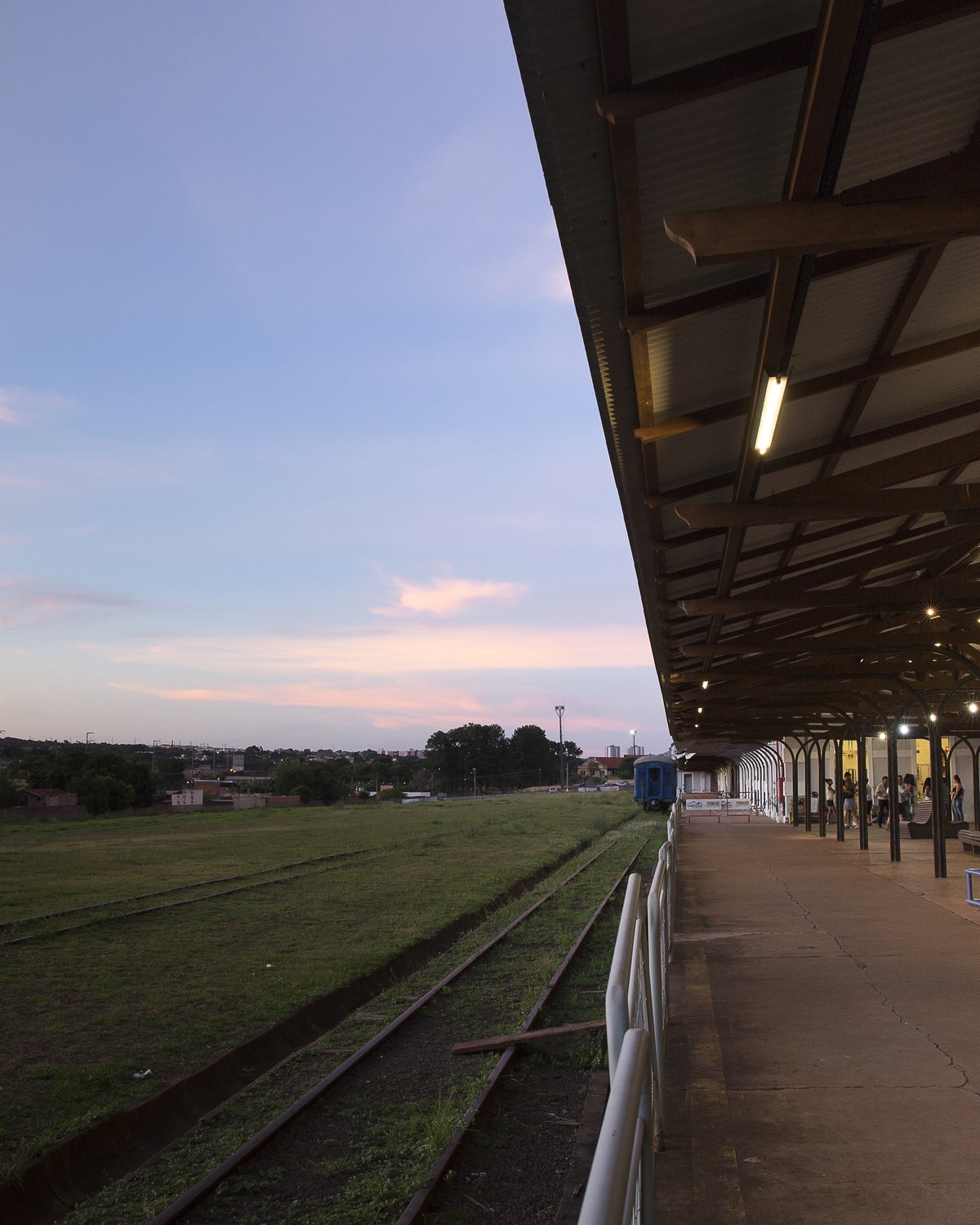 Plataforma coberta da Estação Ferroviária de Campo Grande no fim da tarde, com bancos de ferro, pessoas sentadas, os trilhos ao lado e o céu rosado.