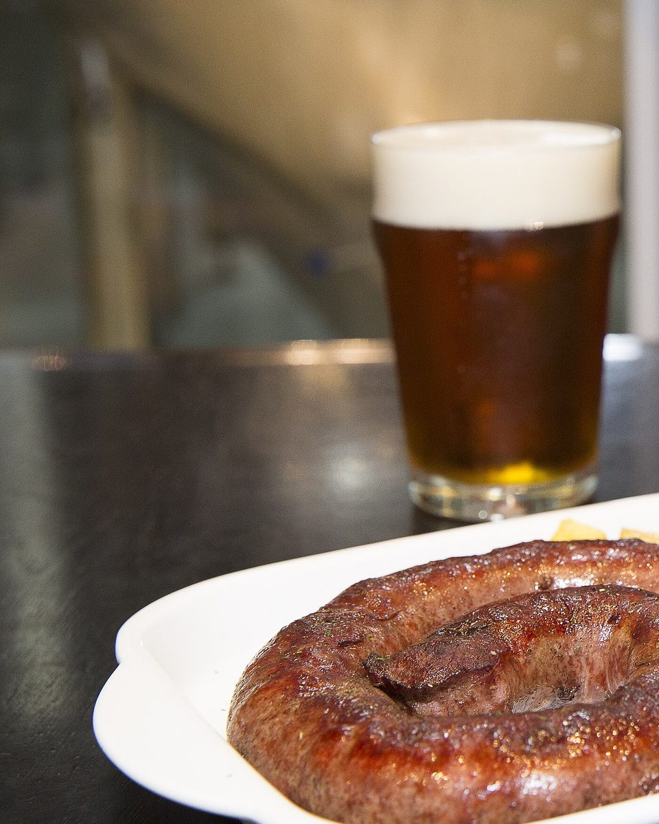 Prato branco com linguiça pantaneira enrolada e mandioca frita, ao lado de um copo de chope, sobre mesa escura numa cervejaria de Campo Grande.