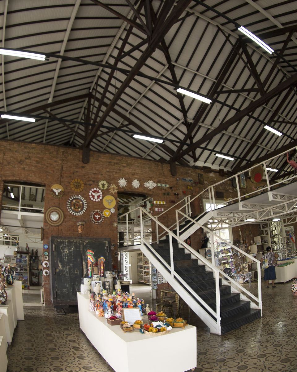 Salão da Casa do Artesão de Campo Grande, com telhado de estrutura de madeira, paredes de tijolo aparente, escada branca e mesas com peças de artesanato à venda.