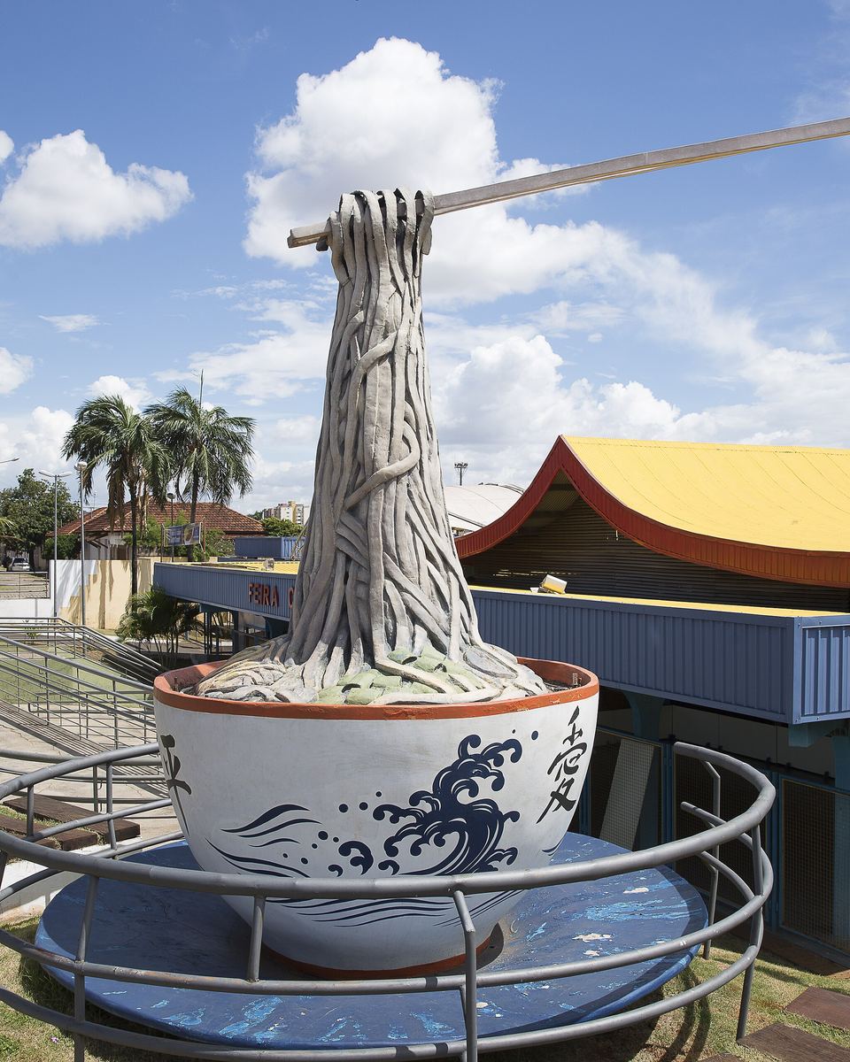 Monumento ao Sobá, uma tigela gigante de concreto com macarrão e um hashi atravessado, instalada em frente ao prédio de telhado amarelo e vermelho da Feira Central.