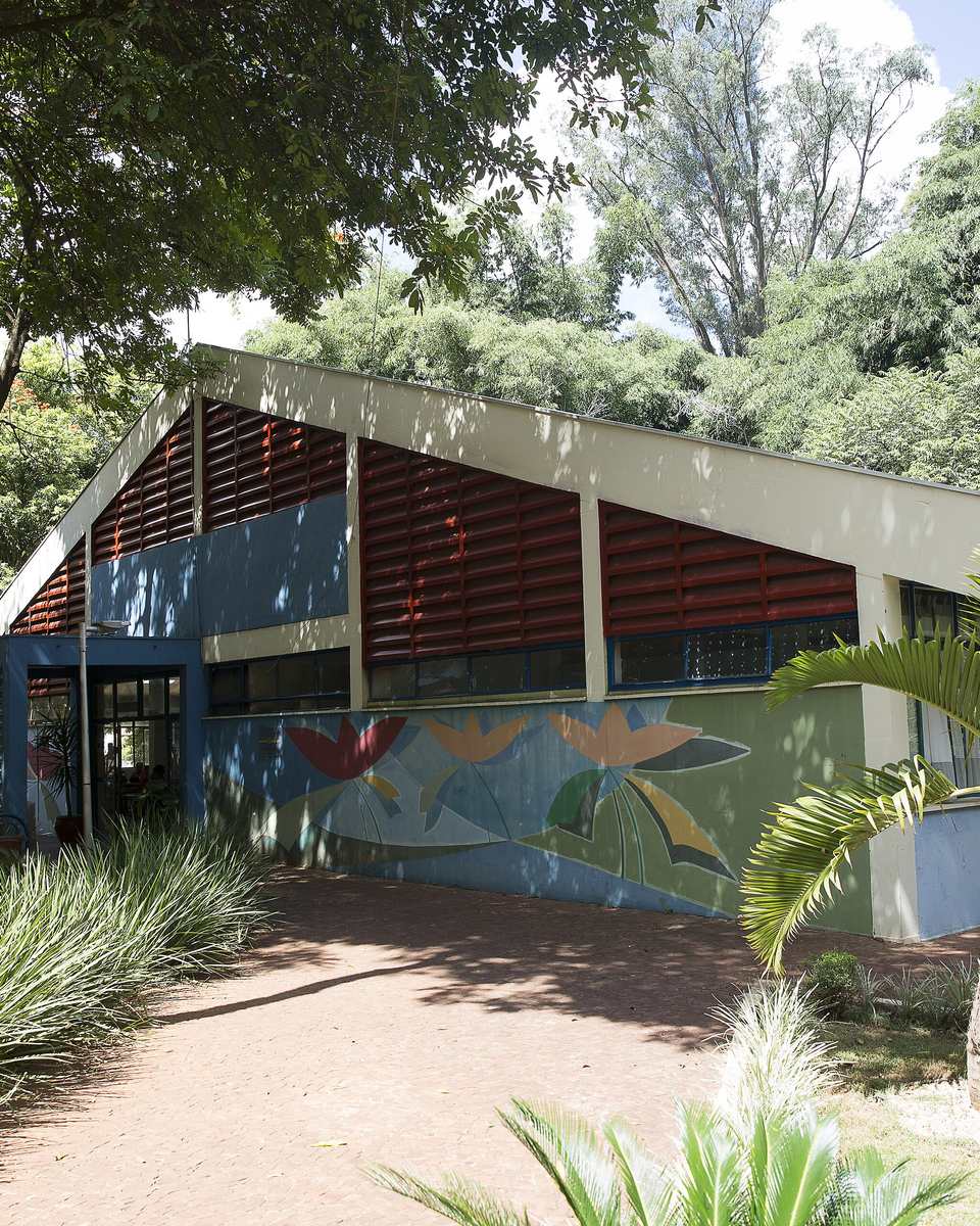Prédio baixo da Biblioteca Municipal de Campo Grande, com brises vermelhos, painel pintado com flores na fachada azul e jardim de palmeiras na frente.