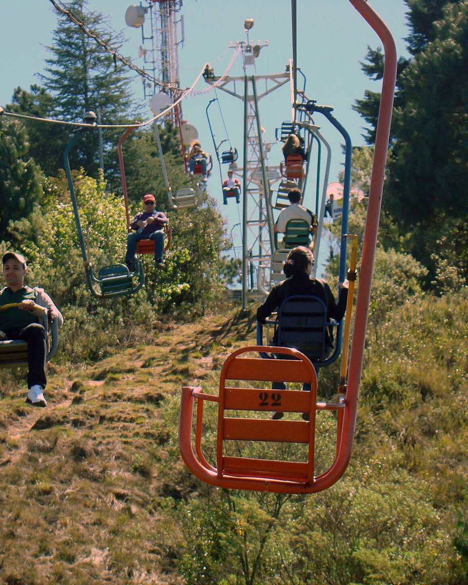 Turistas sentados nas cadeirinhas vermelhas do teleférico de Campos do Jordão, subindo o morro entre árvores e mato seco num dia de sol.