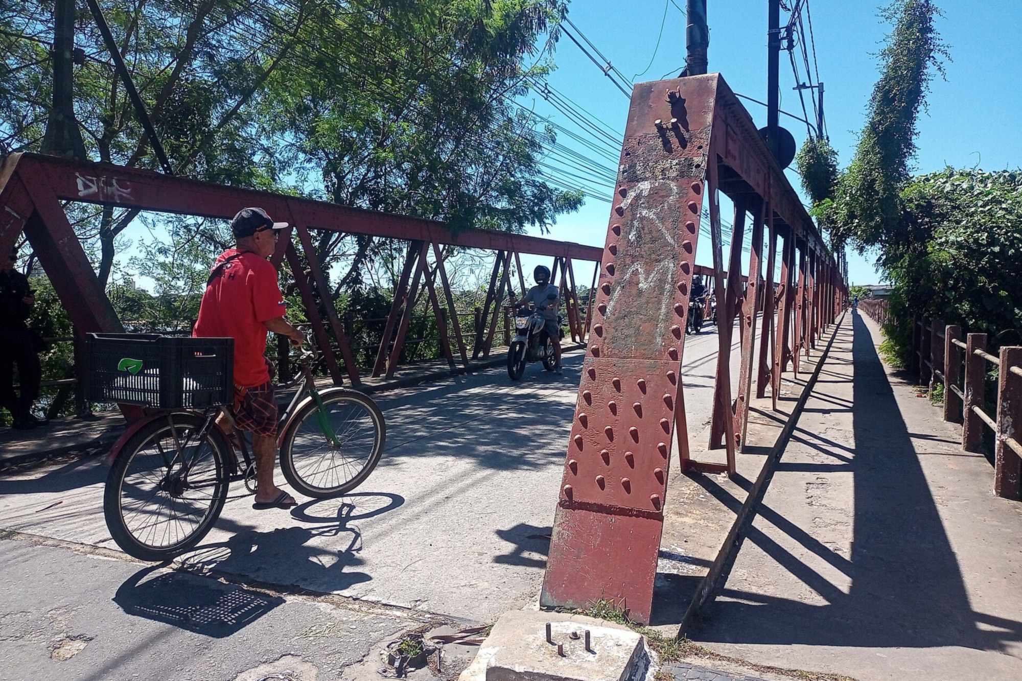 Homem de boné e camiseta vermelha empurrando uma bicicleta com caixote na garupa na entrada da Ponte de Ferro sobre o Rio Paraíba do Sul, no Centro de Campos dos Goytacazes, com um motociclista atravessando a ponte, a estrutura de ferro vermelha e árvores dos dois lados, num dia de sol.