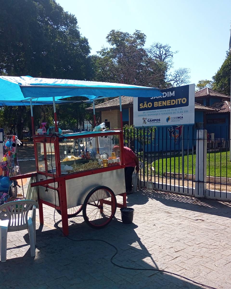 Carrinho de pipoca com toldo azul e um pipoqueiro trabalhando na calçada em frente ao portão do Jardim São Benedito, no Centro de Campos dos Goytacazes, com brinquedos pendurados, uma cadeira de plástico, árvores e um poste vermelho.