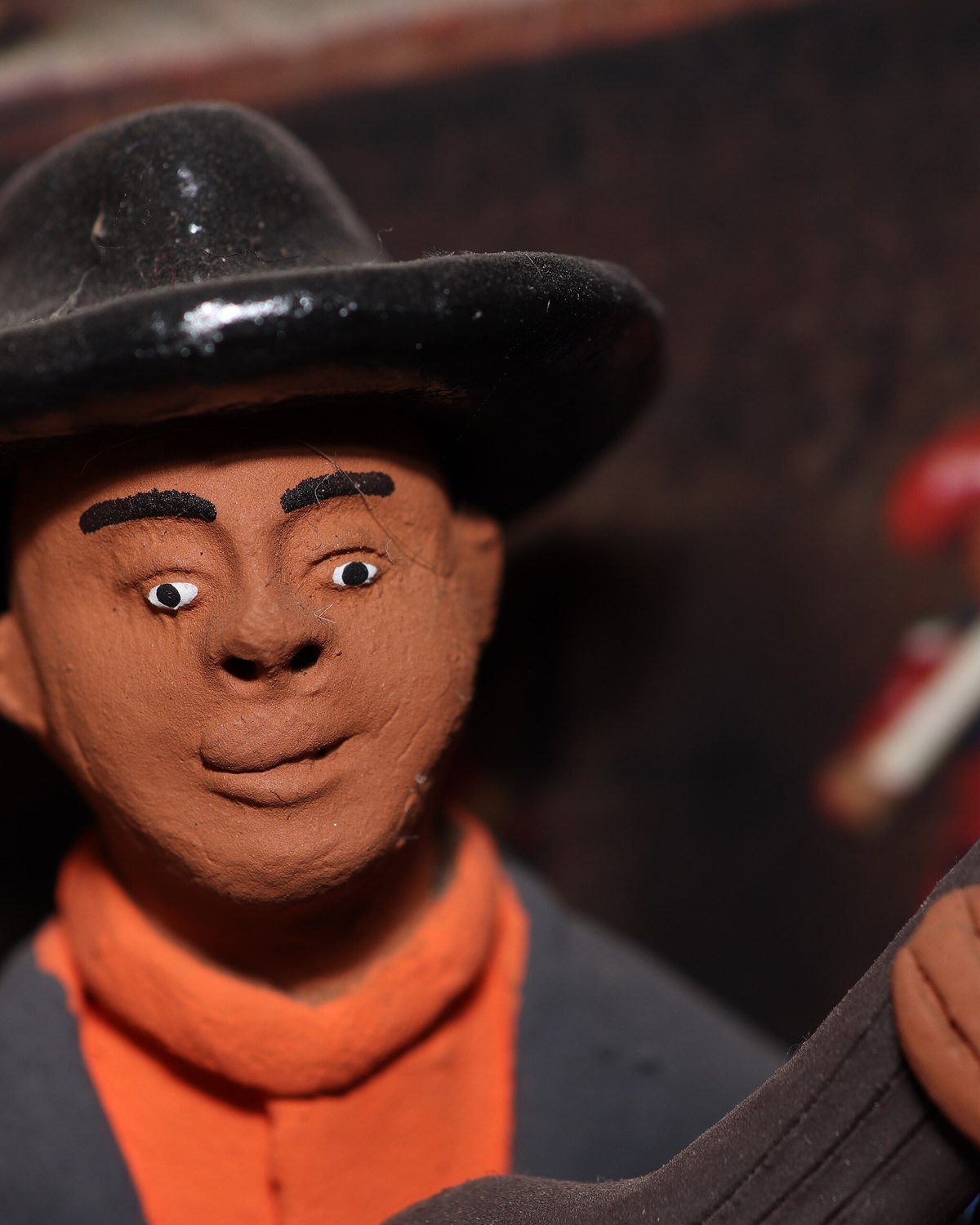 Figura de barro pintada de um violeiro de chapéu preto, lenço laranja e paletó azul, tocando viola, com outras figuras de barro desfocadas ao fundo.