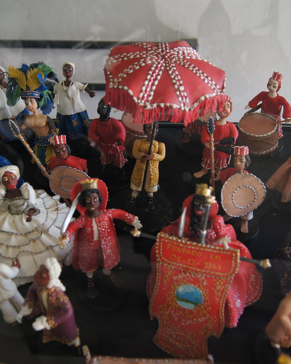 Conjunto de figuras de cerâmica de Caruaru representando um maracatu nação, com rainha e rei sob o pálio vermelho, baianas de saia branca, batuqueiros com tambores e porta-estandarte, visto de cima numa vitrine.