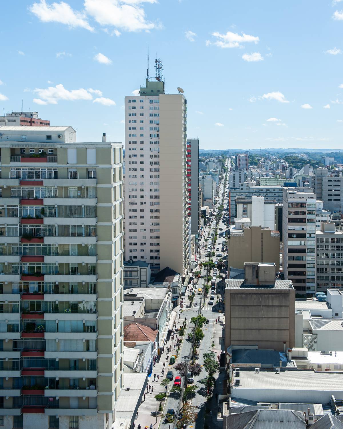 Avenida Júlio de Castilhos vista do alto, em Caxias do Sul, com prédios dos dois lados, árvores no canteiro e carros e pedestres na rua do comércio, num dia de sol.