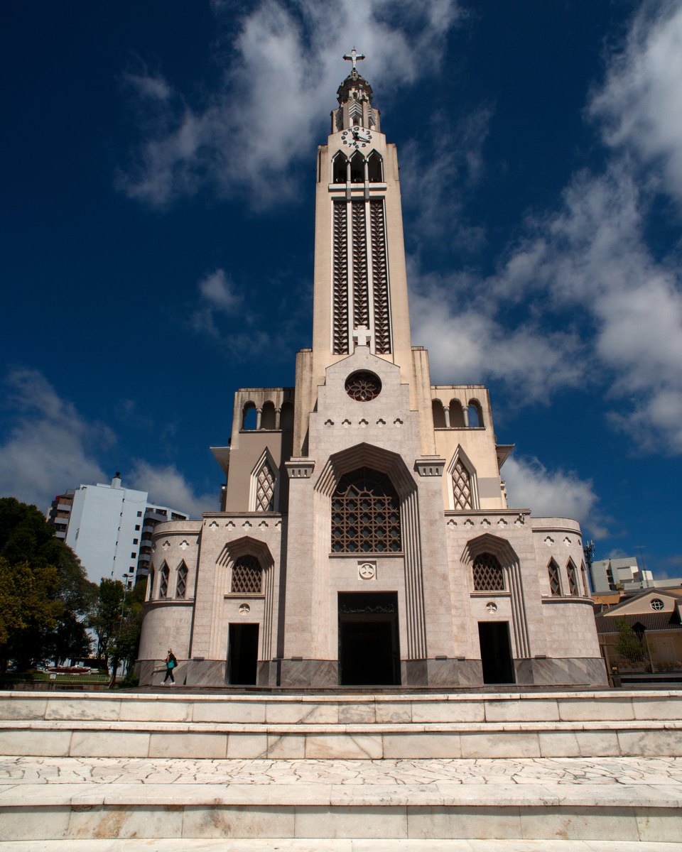 Fachada branca da Igreja de São Pelegrino, em Caxias do Sul, com a torre alta contra o céu azul com nuvens e a escadaria de pedra em primeiro plano.