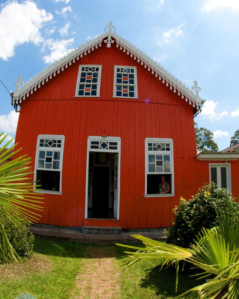 Casa de madeira pintada de vermelho, com janelas brancas e lambrequins no telhado, cercada de gramado e palmeiras numa fazenda de Criúva, no interior de Caxias do Sul, sob céu azul.