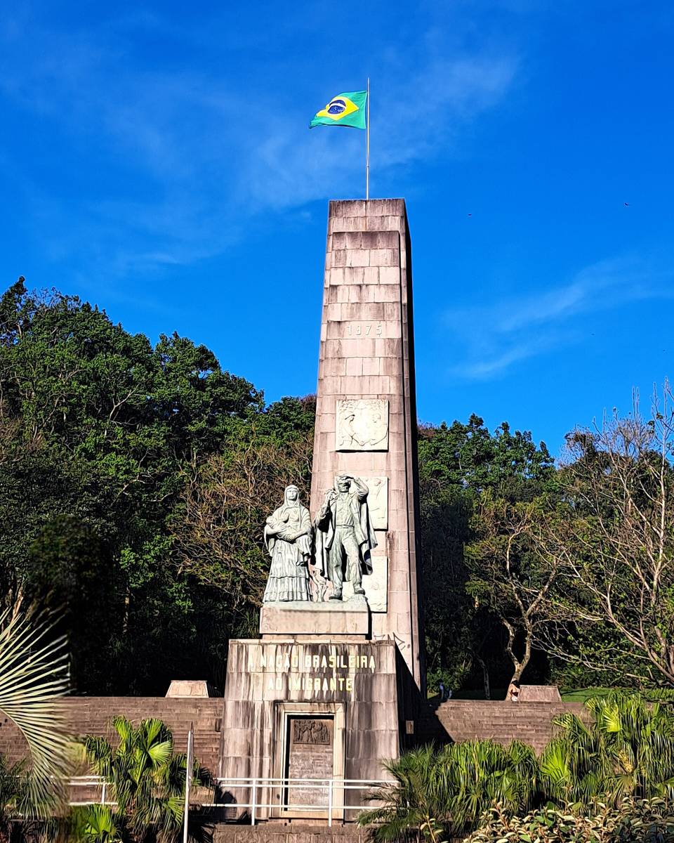 Monumento Nacional ao Imigrante, em Caxias do Sul, com a torre de pedra, a bandeira do Brasil no alto e as estátuas de um casal de imigrantes na base, entre árvores, sob céu azul.