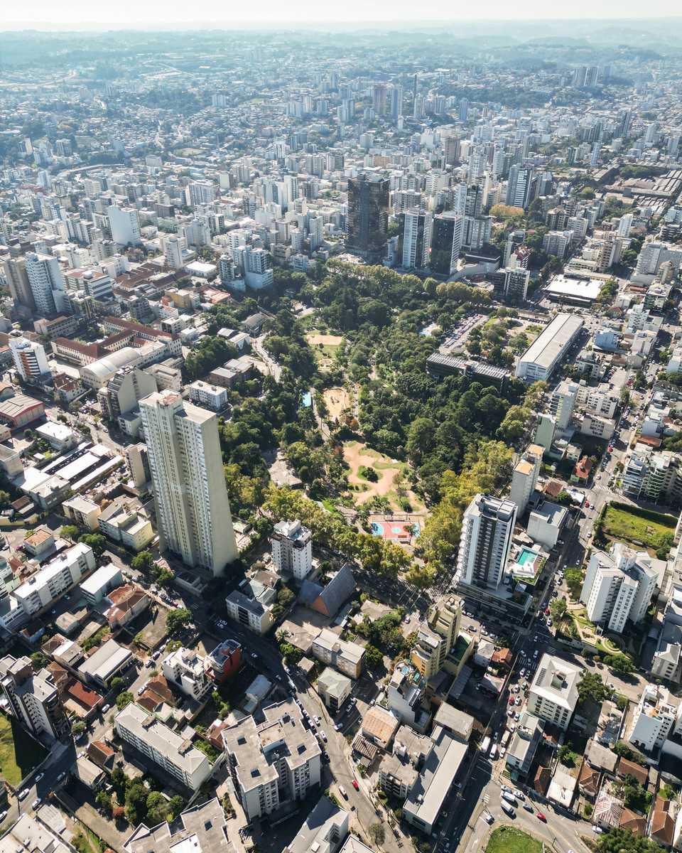 Vista aérea vertical do centro de Caxias do Sul, com prédios altos, ruas em diagonal e um parque arborizado no meio da cidade, sob céu claro.