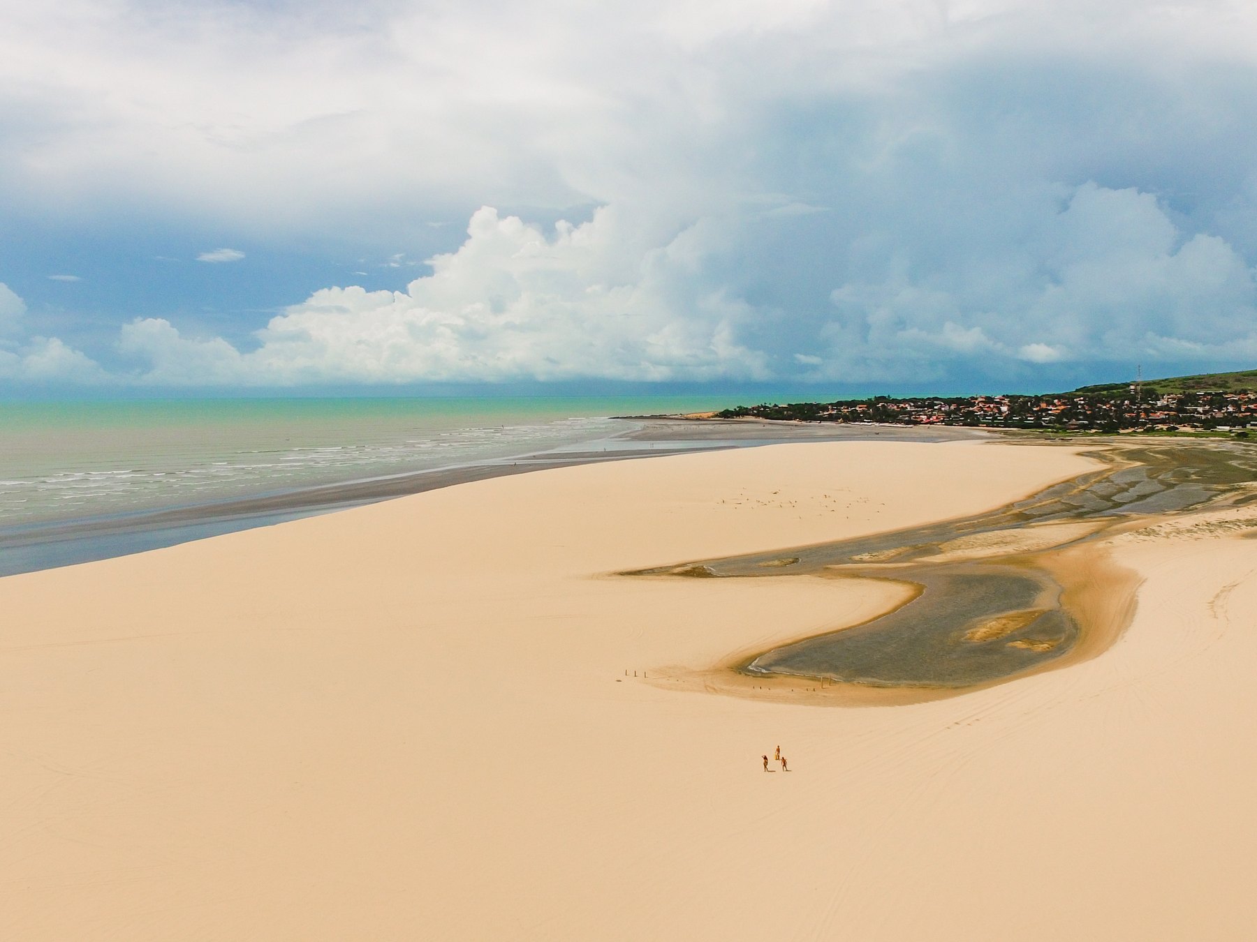 Vista aérea da Duna do Pôr do Sol, em Jericoacoara, com a areia clara em curva, uma lagoa rasa se formando ao pé da duna, duas pessoas caminhando e o mar verde-azulado com a vila ao fundo sob nuvens altas.