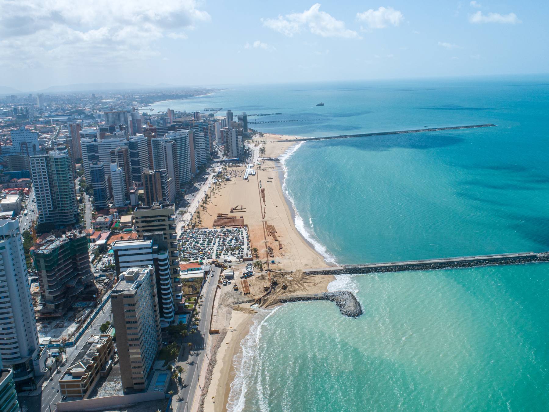 Vista aérea da orla da Beira-Mar, em Fortaleza, com os prédios à esquerda, a faixa de areia, os espigões avançando no mar e a água azul-turquesa até o horizonte.