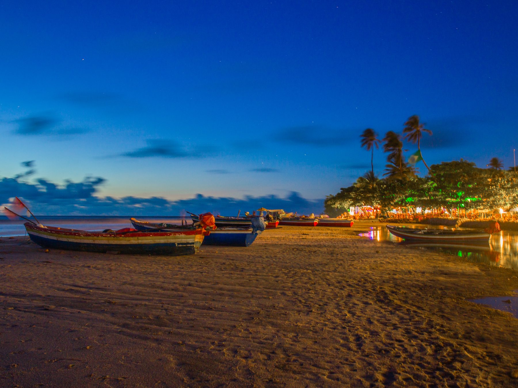 Praia de Jericoacoara ao anoitecer, com barcos de pesca na areia molhada, o céu azul-escuro e as luzes dos bares e restaurantes da vila acesas entre coqueiros.