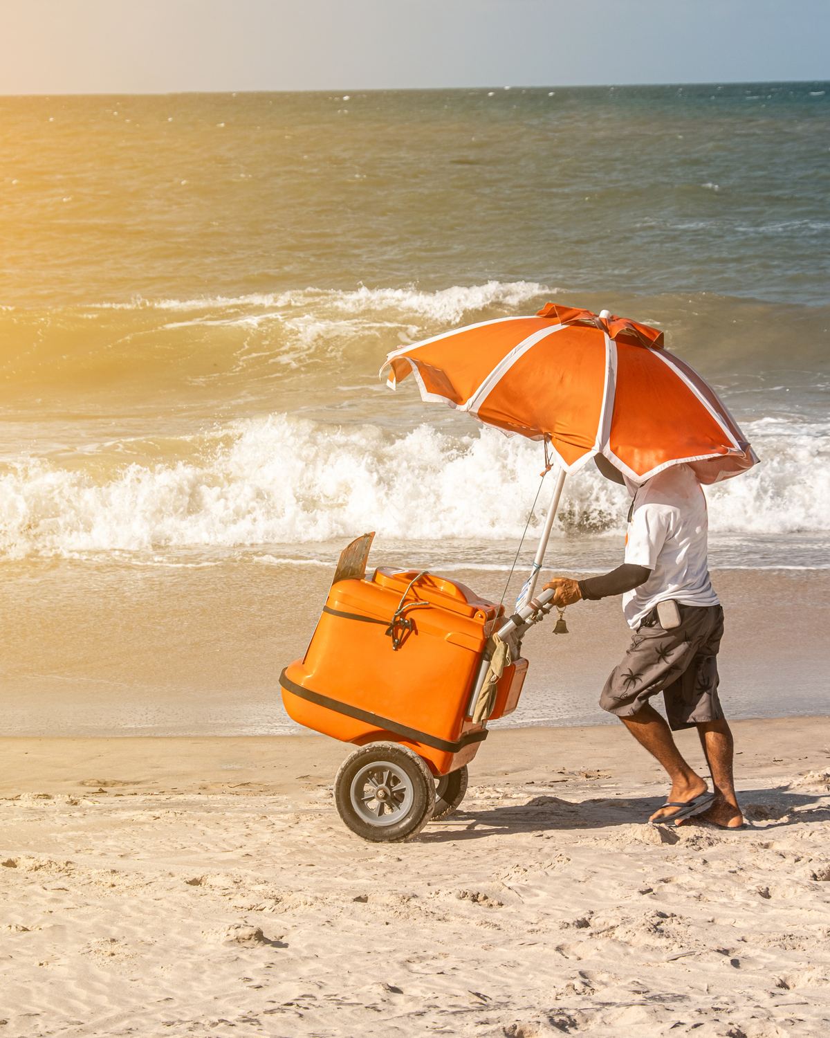 Vendedor ambulante de camiseta branca e bermuda empurrando um carrinho com caixa térmica laranja e guarda-sol pela areia do Cumbuco, com as ondas atrás na luz dourada da tarde.