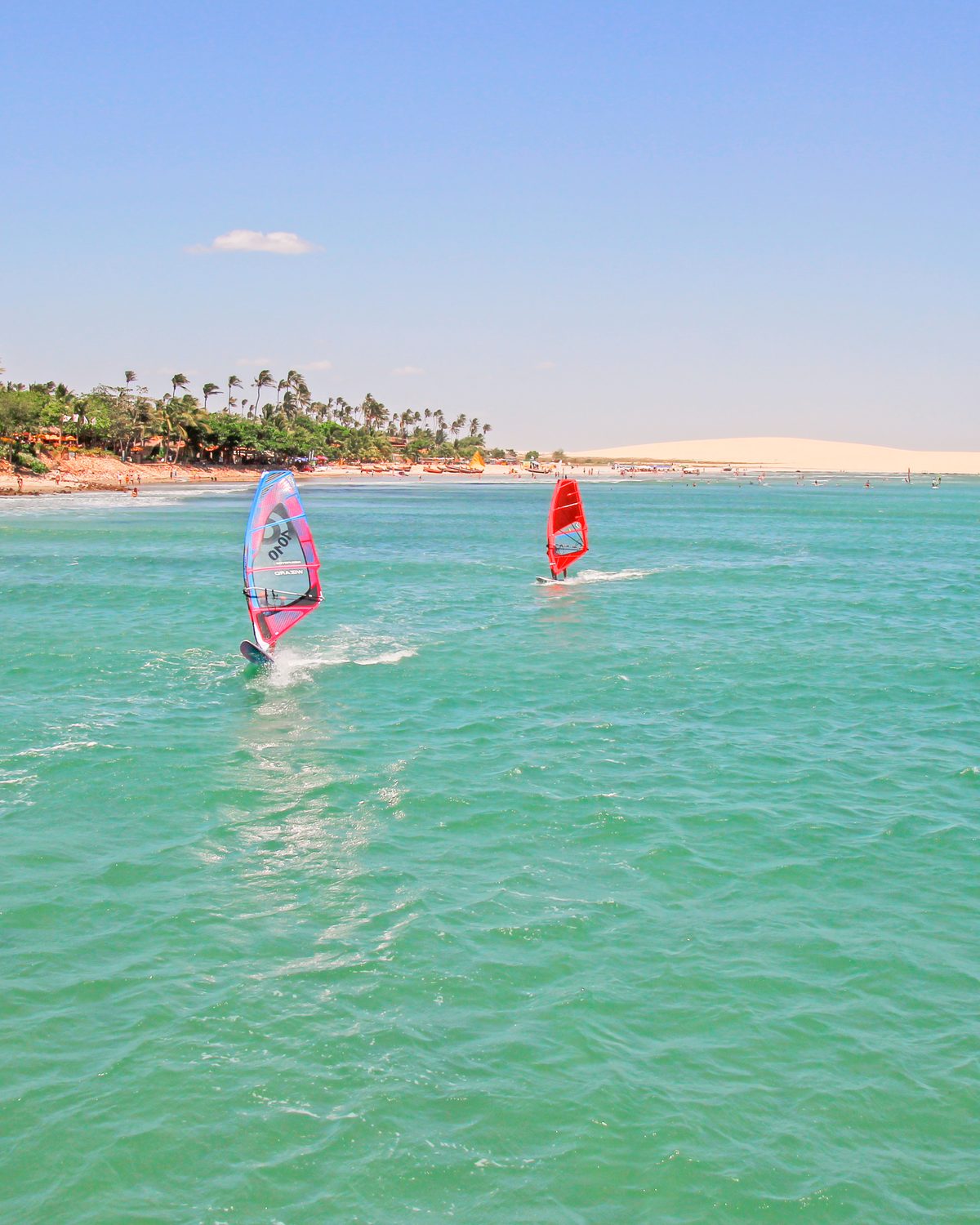 Dois windsurfistas velejando no mar verde-turquesa de Jericoacoara, com barrancos avermelhados, coqueiros e dunas claras ao fundo.