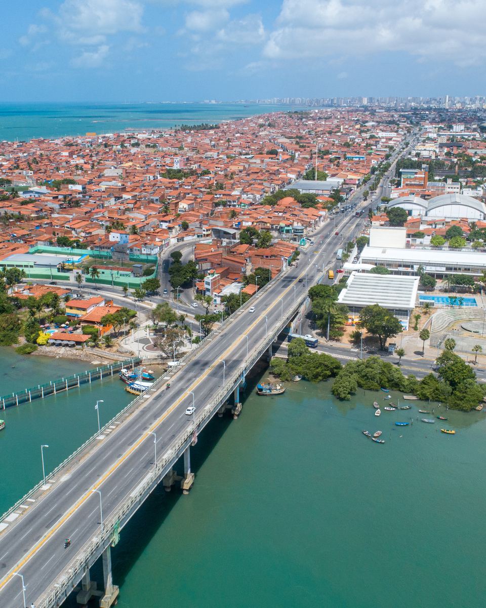Vista aérea da ponte sobre o Rio Ceará, na divisa entre Fortaleza e Caucaia, com o rio verde, barcos atracados e a cidade espalhada até o mar no horizonte.