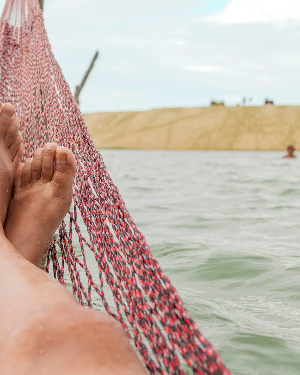 Pés de uma pessoa deitada numa rede vermelha armada dentro da Lagoa de Tatajuba, perto de Jericoacoara, com a água verde, banhistas e a duna ao fundo.