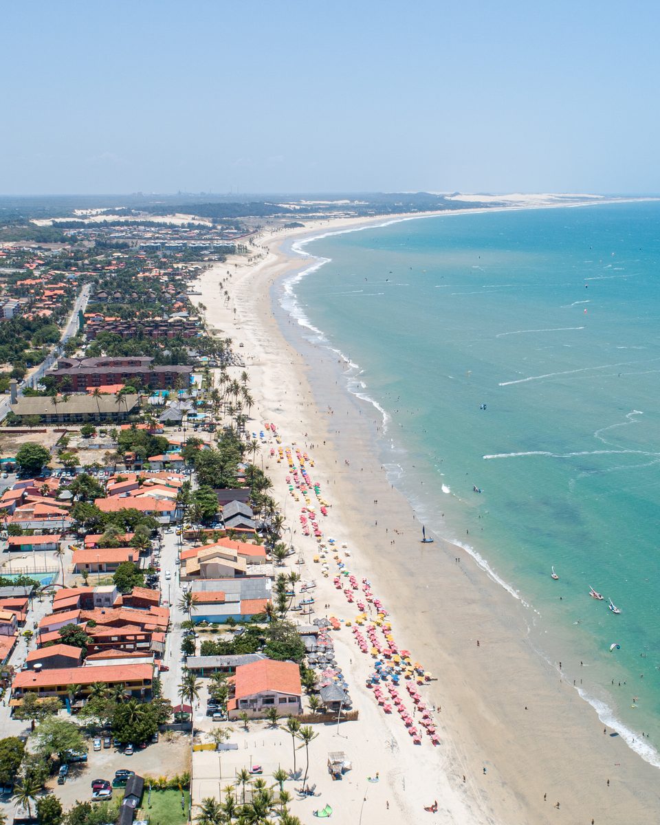 Vista aérea da Praia do Cumbuco, em Caucaia, com a vila de telhados vermelhos, a longa faixa de areia com barracas e o mar azul-turquesa pontilhado de kitesurfistas.