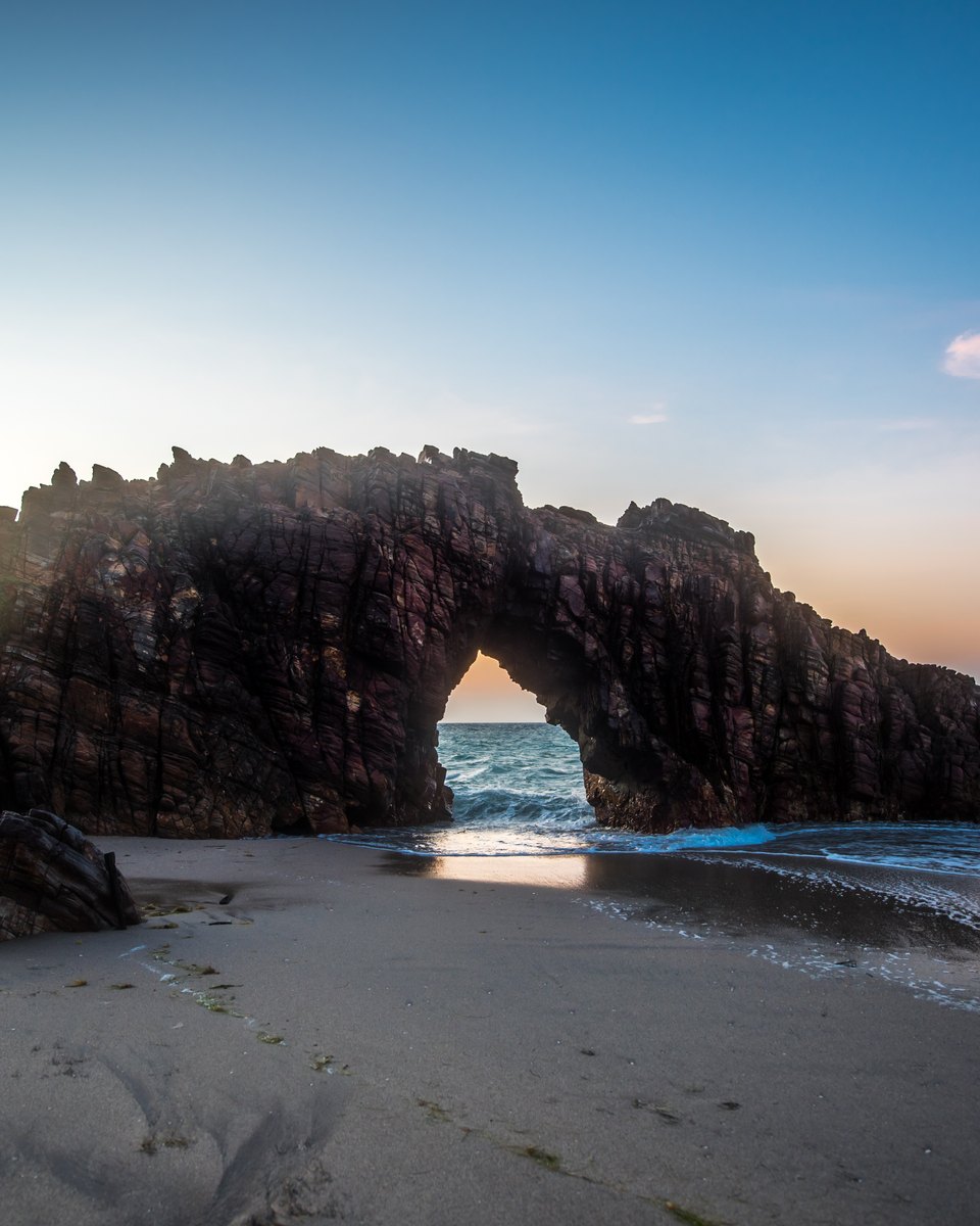 Pedra Furada de Jericoacoara ao pôr do sol, um arco de rocha escura sobre a areia molhada, com o sol baixo aparecendo pela abertura e o céu azul com nuvens rosadas.