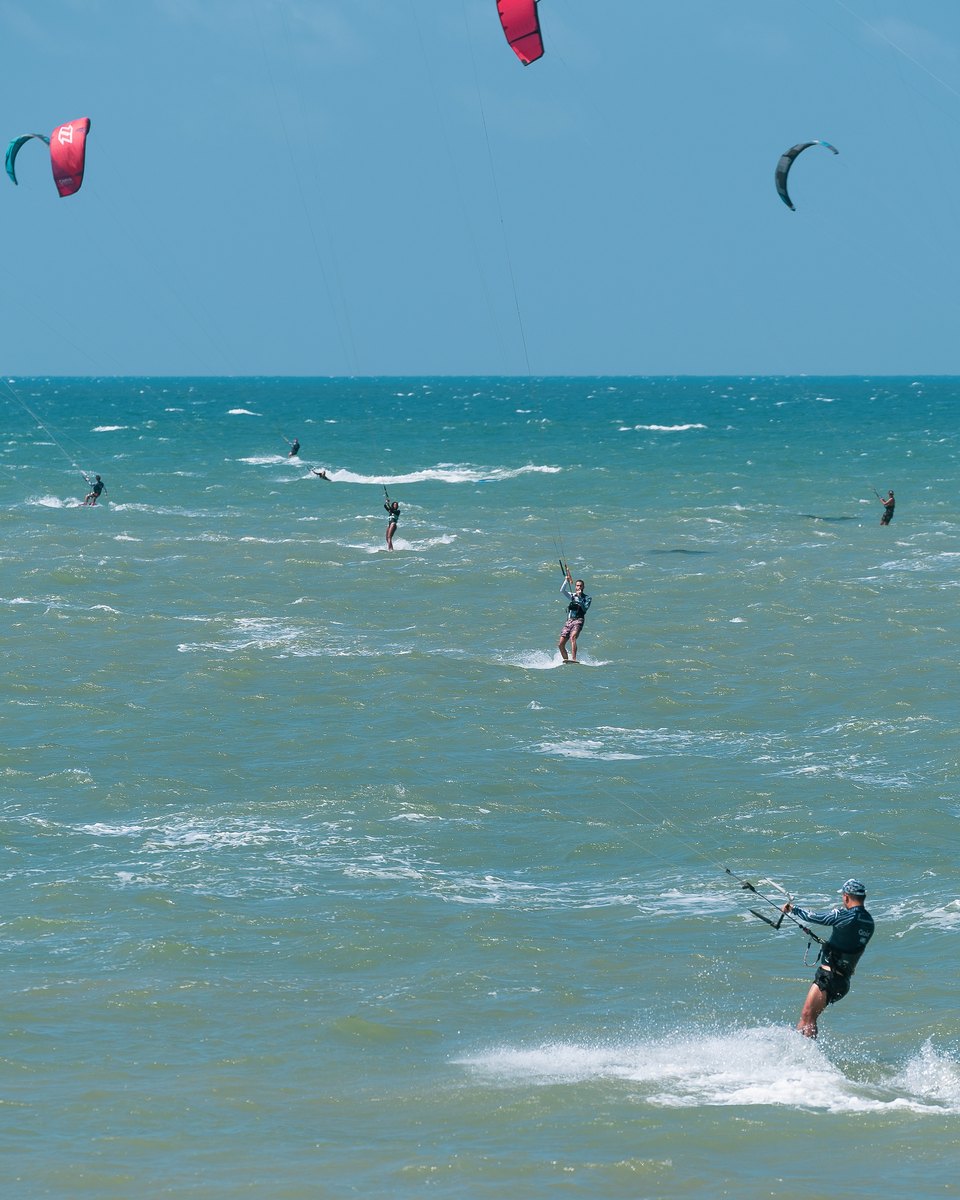 Dezenas de kitesurfistas no mar azul da Praia do Cumbuco, com as pipas coloridas no céu e um velejador em primeiro plano levantando água.