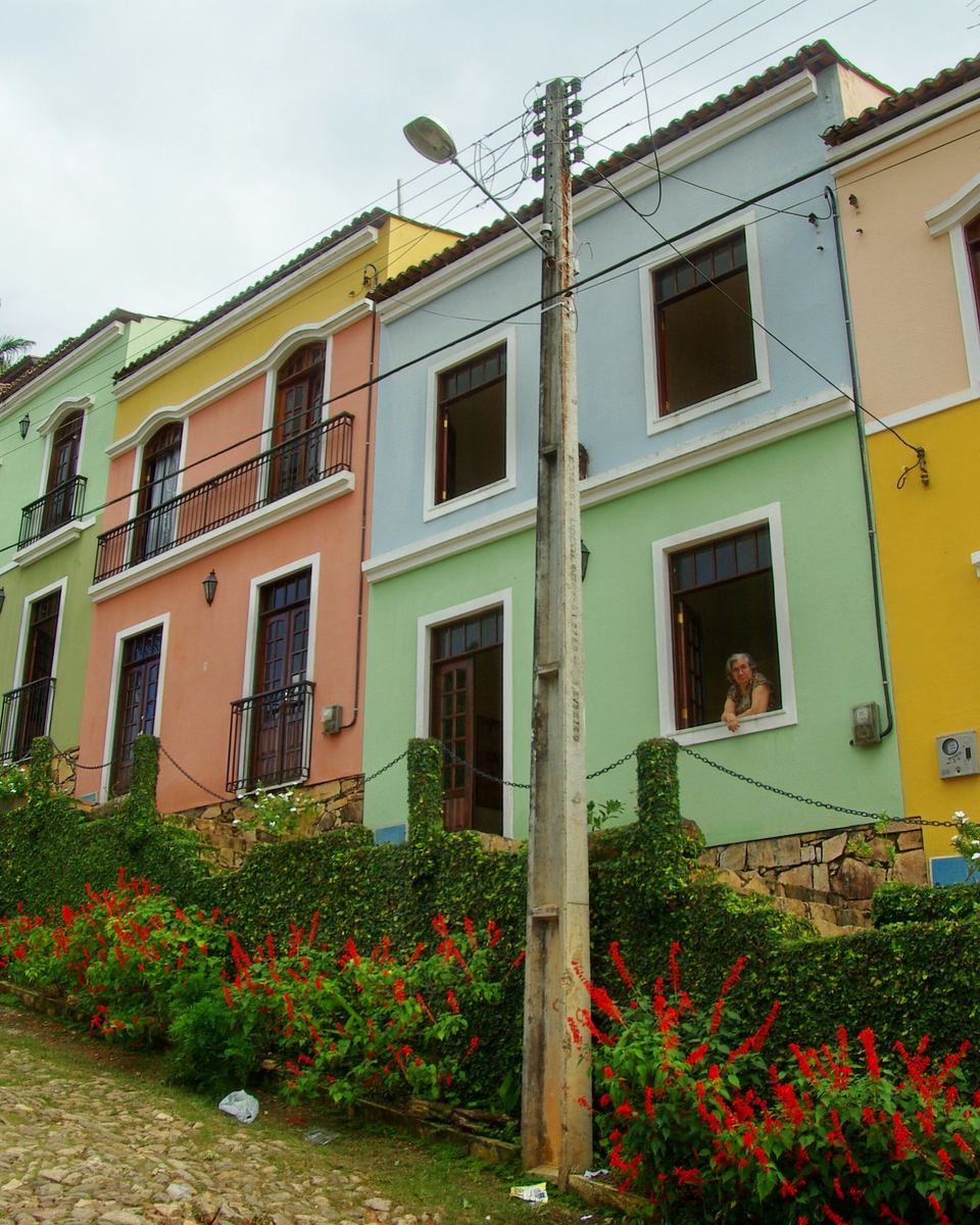 Rua de pedra em Guaramiranga com uma fileira de casas coloridas, amarela, verde, rosa e salmão, e canteiros de flores vermelhas na frente.