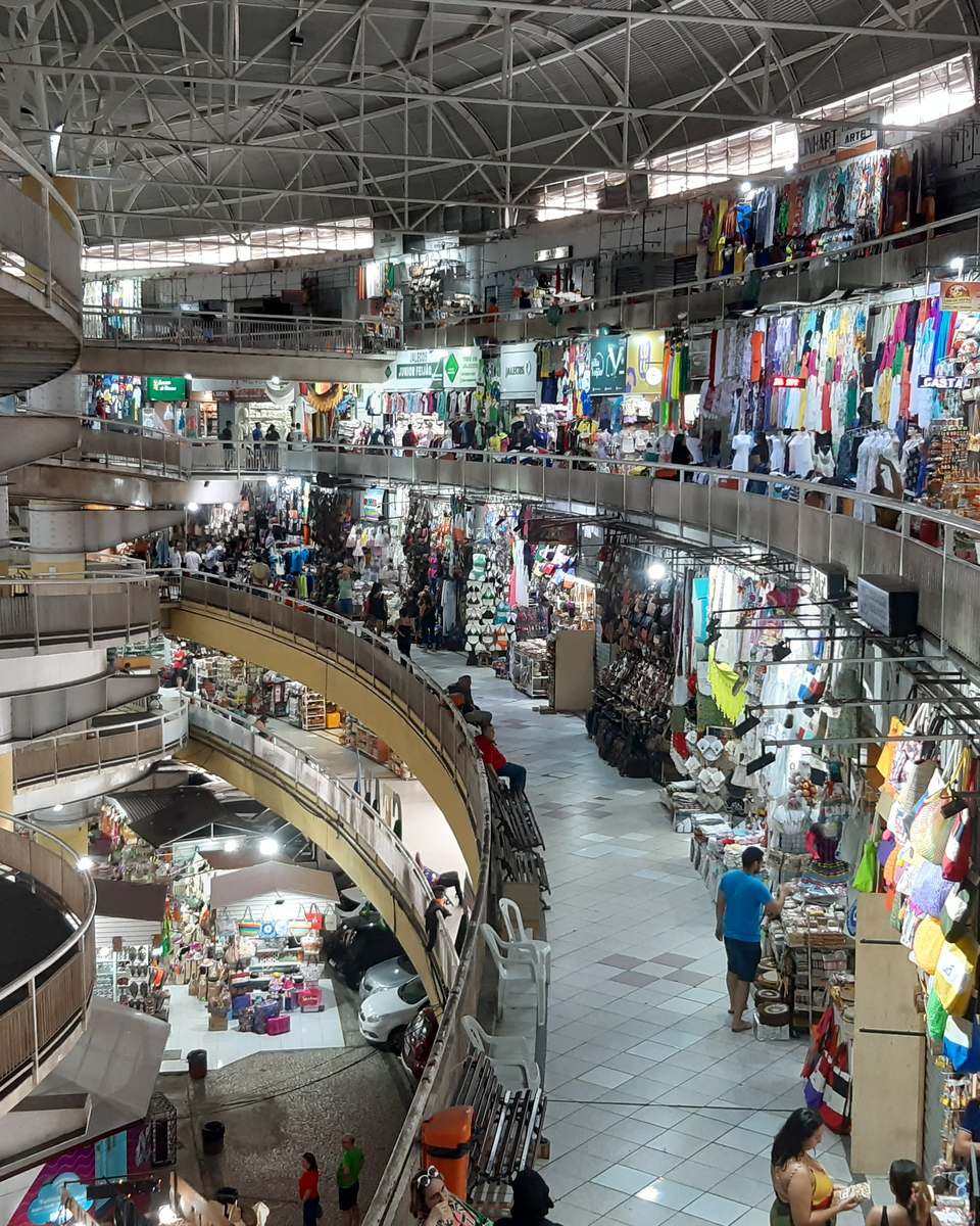 Interior do Mercado Central de Fortaleza visto do alto, com os andares em curva cheios de boxes de roupas, redes e artesanato coloridos, rampas e pessoas circulando.