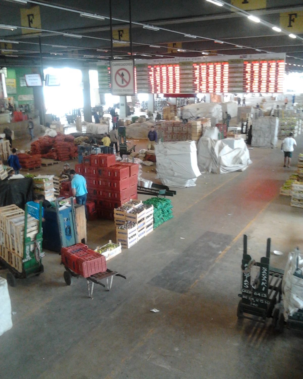 Pavilhão da CeasaMinas, em Contagem, com caixas de frutas e verduras empilhadas, carrinhos de carga e trabalhadores circulando entre as bancas sob os painéis de preço.