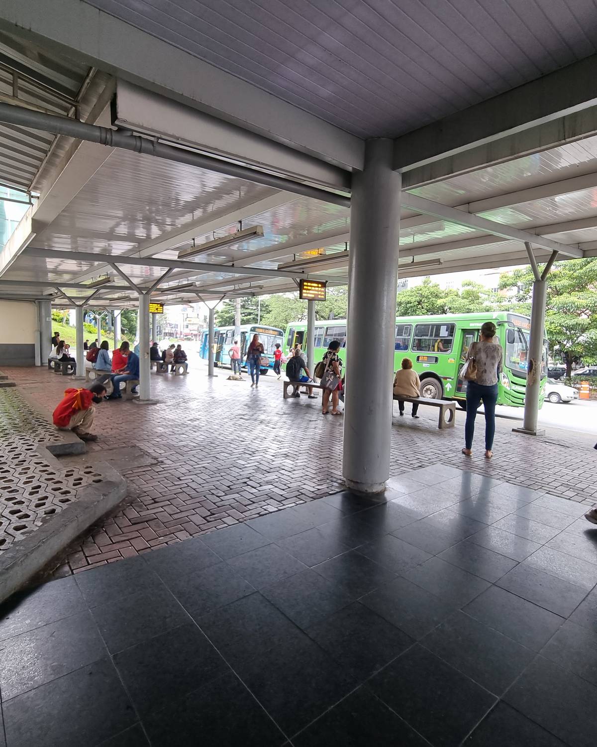 Duas mulheres caminhando de braços dados no ponto de ônibus coberto em frente ao Big Shopping, no Eldorado, com passageiros esperando nos bancos e um ônibus verde parado ao fundo.