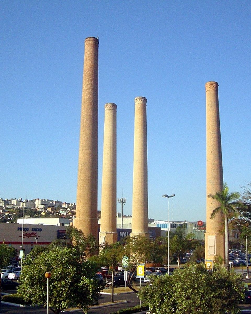 As quatro chaminés de tijolo da antiga fábrica de cimento Itaú, altas e finas, entre palmeiras e postes no estacionamento do ItaúPower Shopping, em Contagem, com um morro de casas ao fundo sob céu azul.