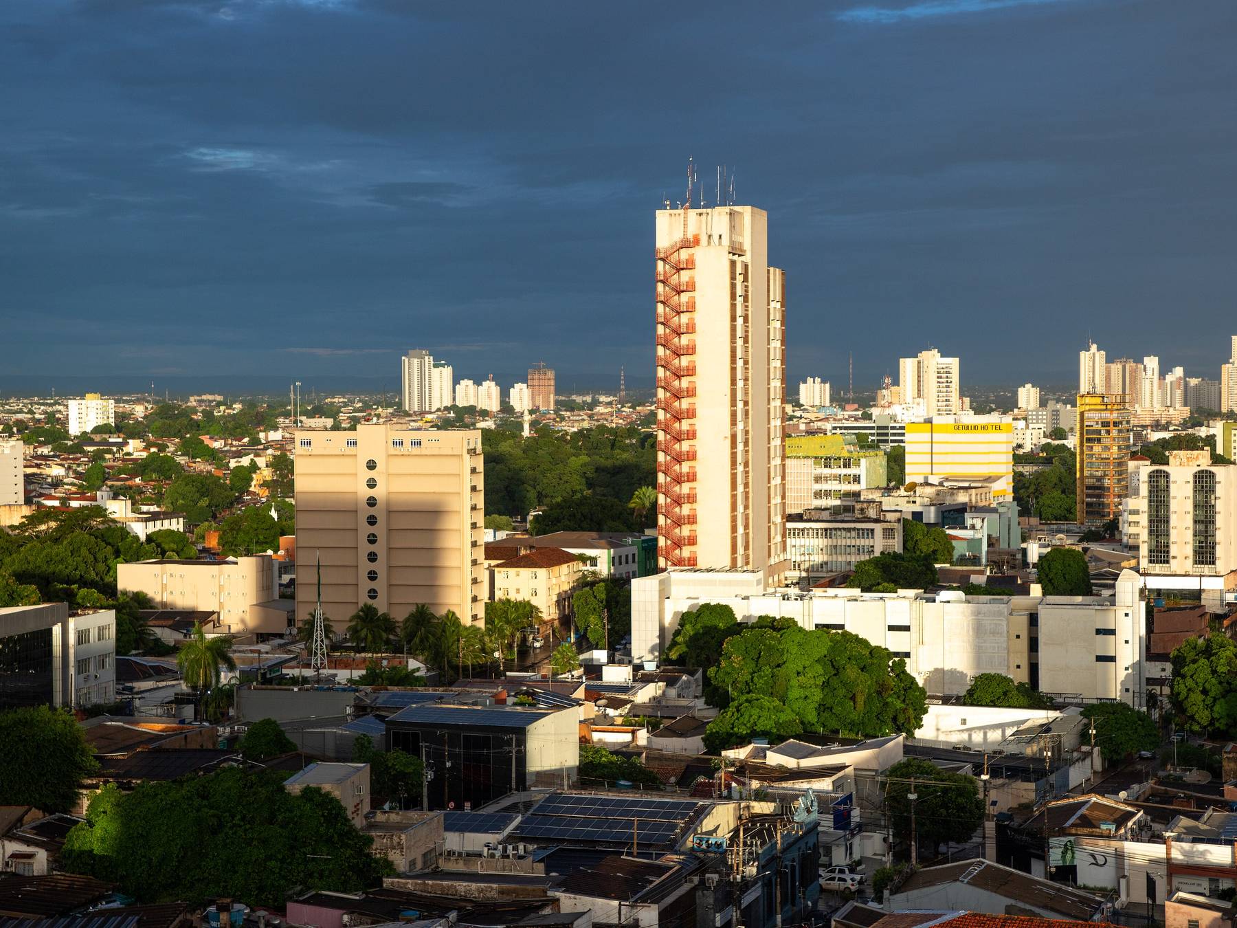 Prédios do centro de Cuiabá vistos do alto no fim da tarde, com a luz dourada batendo nas torres, as copas verdes das árvores entre os quarteirões e um céu carregado de chuva ao fundo