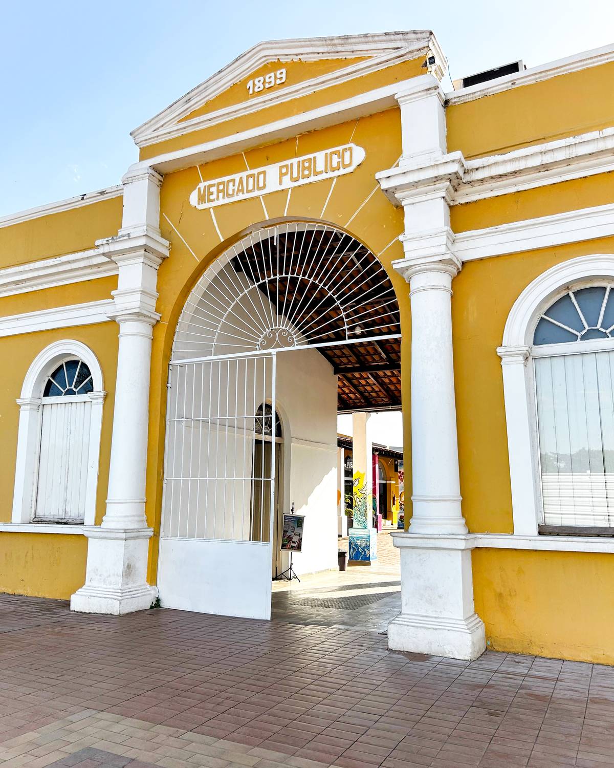 Fachada amarela do Mercado do Peixe, no bairro do Porto, com o portão de ferro branco em arco, colunas brancas e a data de 1899 no frontão.