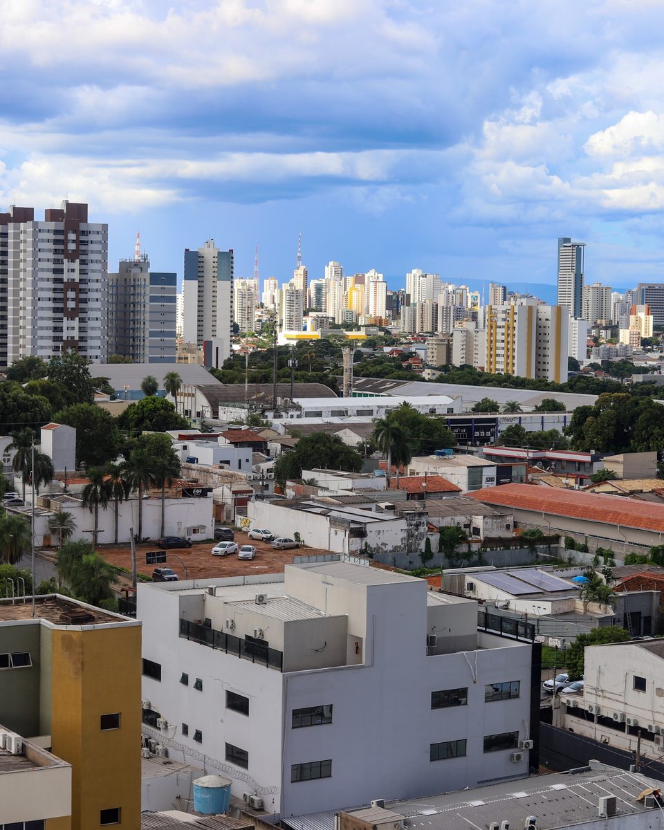 Cuiabá vista do alto, com prédios altos no horizonte, telhados de barro e árvores no primeiro plano e nuvens carregadas no céu.
