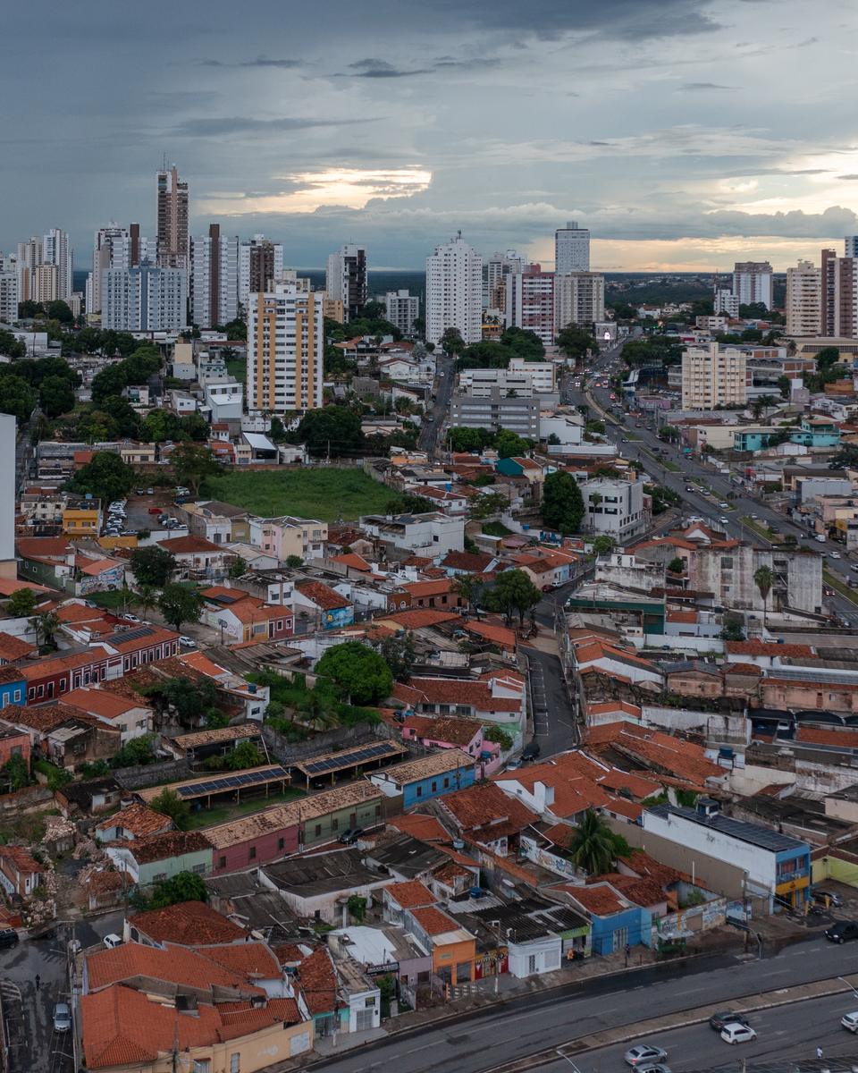 Centro Histórico de Cuiabá visto do alto no fim da tarde, com telhados de barro das casas antigas em primeiro plano e os prédios do centro novo ao fundo.
