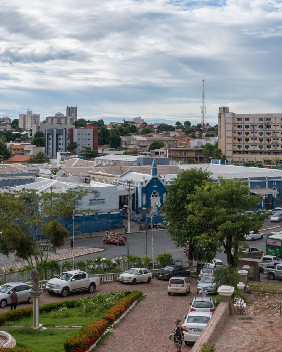Centro de Cuiabá visto do Morro do Seminário, com carros estacionados, um ônibus na avenida, lojas de fachada azul e prédios ao fundo.