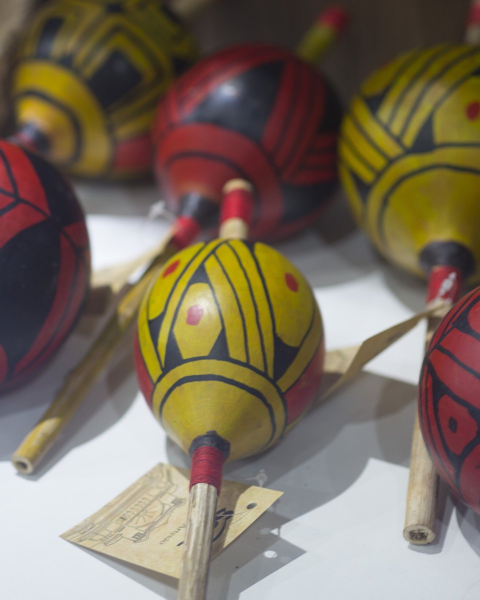 Ganzás de cabaça pintados de vermelho, amarelo e preto, expostos lado a lado sobre uma bancada na Casa do Artesão, em Cuiabá.