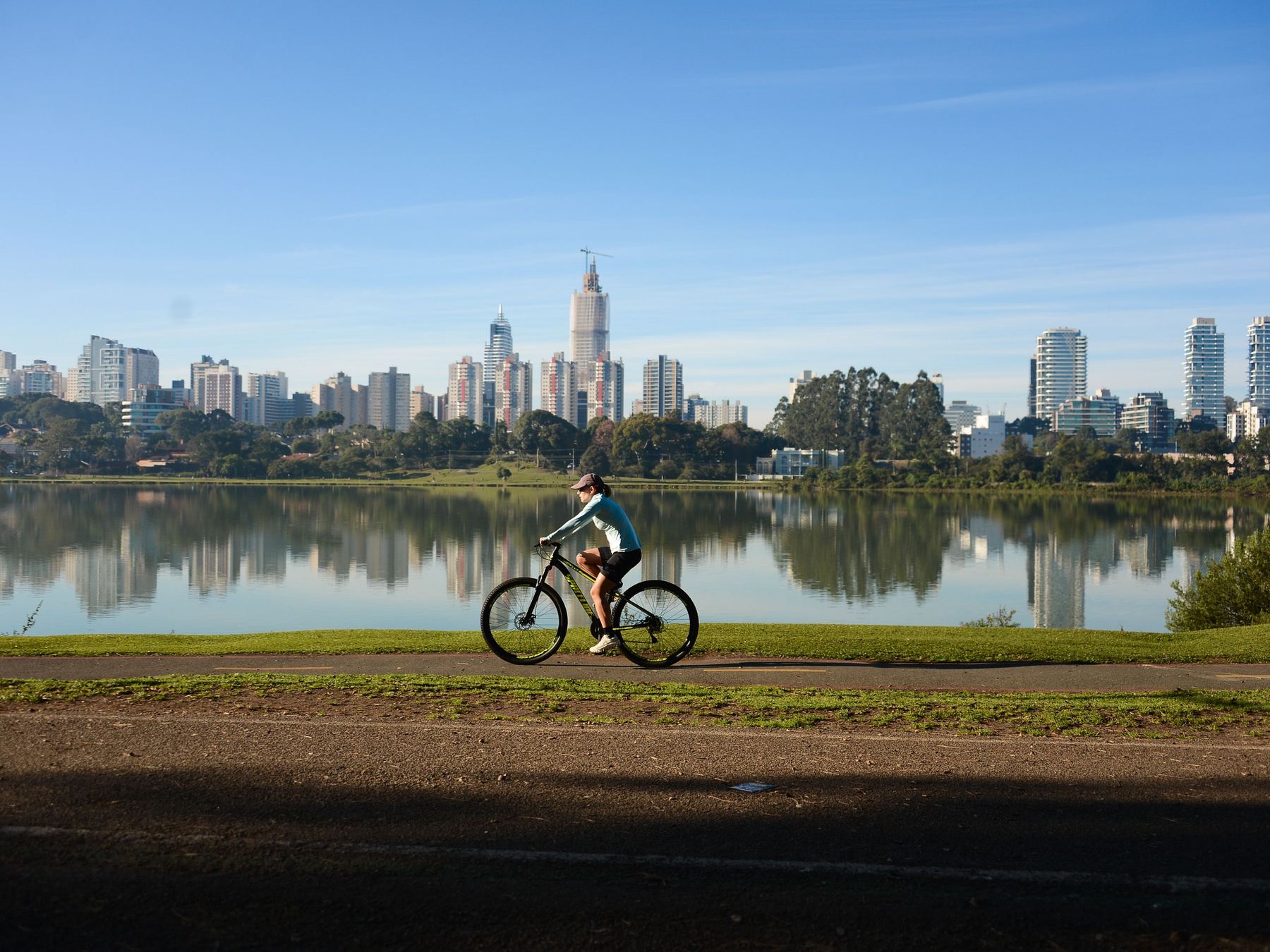 Ciclista pedalando na margem do lago do Parque Barigui num dia de céu azul, com os prédios de Curitiba refletidos na água ao fundo.