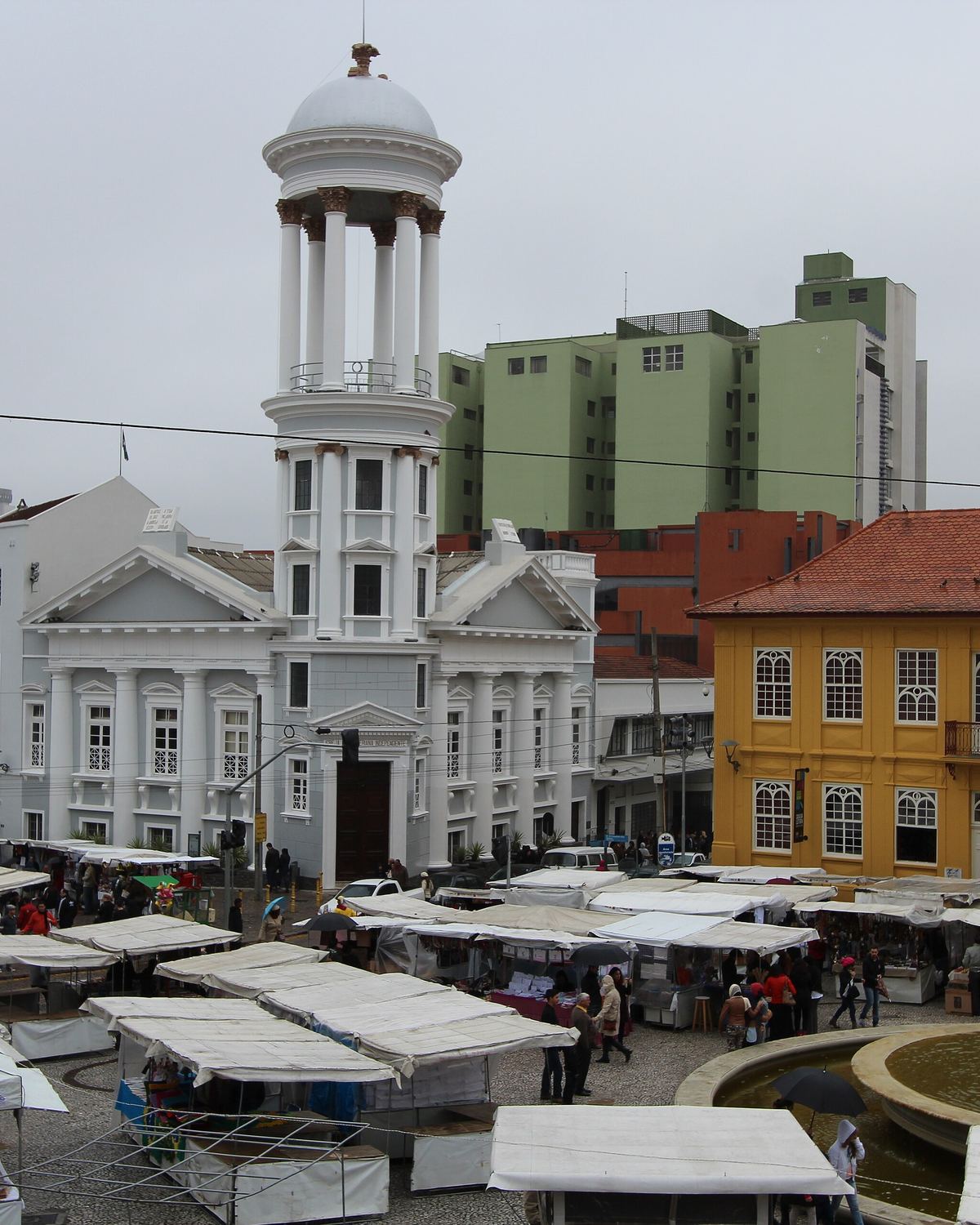 Barracas brancas da feira de artesanato de domingo no Largo da Ordem, no centro histórico de Curitiba, vistas do alto, com a igreja de torre branca e os casarões coloridos ao fundo.