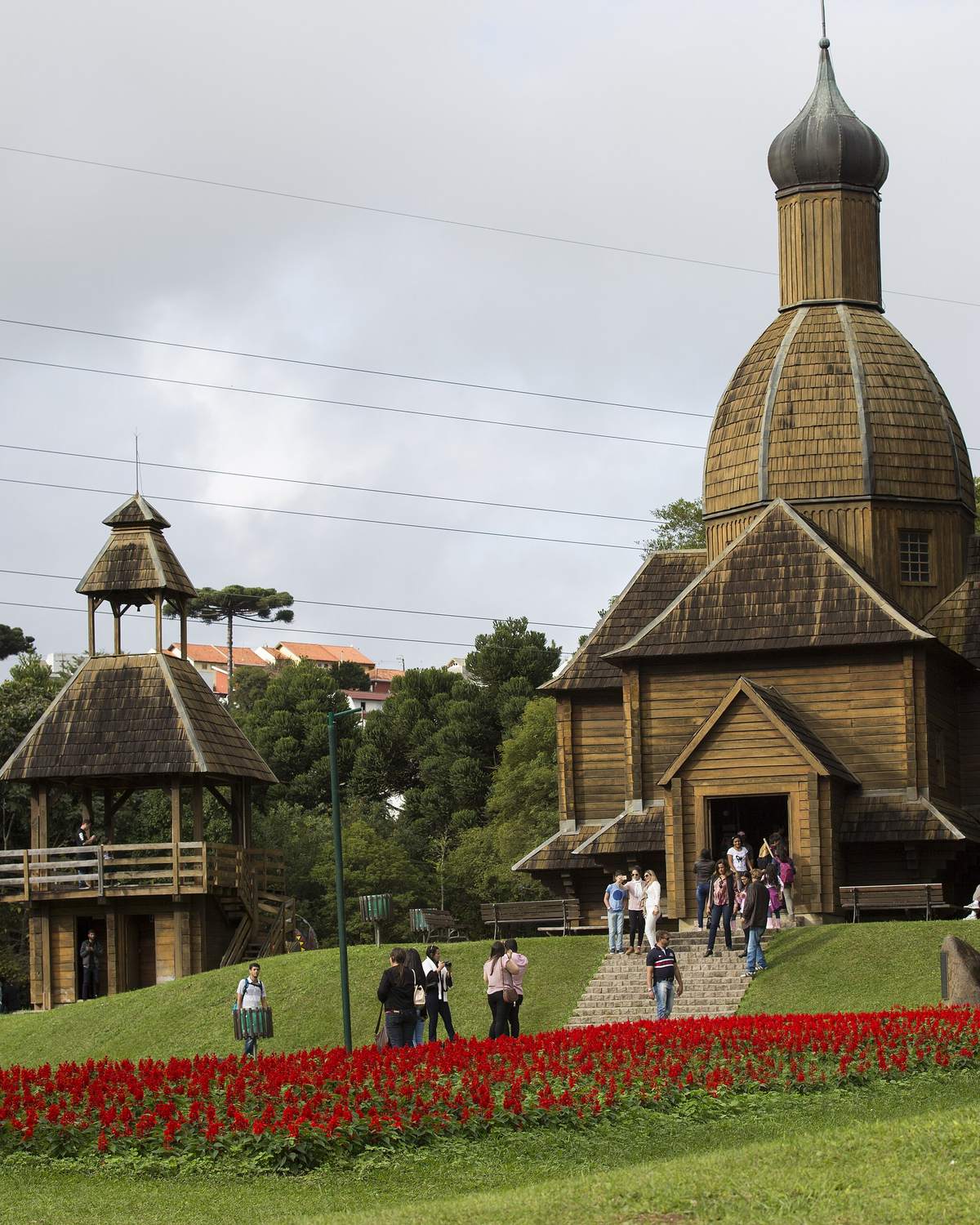 Visitantes no gramado do Parque Tingui, em Curitiba, diante da igreja de madeira com cúpula do Memorial Ucraniano, cercada de araucárias.