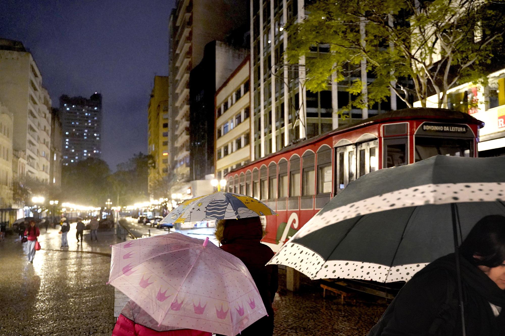 Pedestres com guarda-chuvas caminhando à noite pelo calçadão molhado da Rua XV de Novembro, em Curitiba, ao lado do bondinho vermelho e das vitrines iluminadas, num dia de garoa.